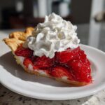 A close-up of a slice of strawberry pie topped generously with whipped cream, showcasing the bright red filling.