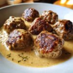 Close-up of several browned Swedish meatballs submerged in a rich, creamy brown sauce, garnished with herbs.