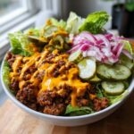 A close-up of a hearty cheeseburger bowl featuring seasoned ground beef, melted cheese sauce, crisp lettuce, pickles, and red onions.