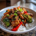 A close-up of a bowl of teriyaki chicken stir fry served over white rice, garnished with sesame seeds and green onions.