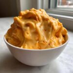 A close-up of a generous serving of thick, bright orange beer cheese dip piled high in a white bowl.
