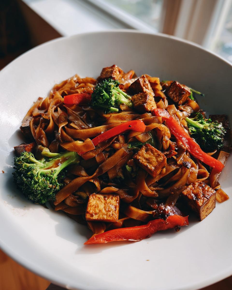A white bowl filled with glossy, dark brown vegetarian drunken noodles, mixed with crispy tofu cubes, bright green broccoli, and red bell peppers.
