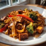 A close-up of a plate of savory vegetarian drunken noodles mixed with pan-fried tofu cubes, bright green broccoli florets, and red bell pepper strips.