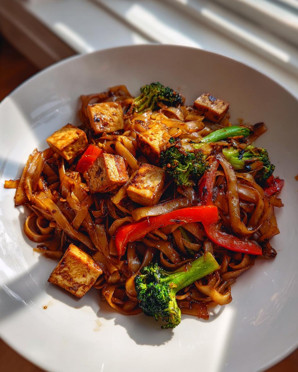 A white bowl filled with saucy vegetarian drunken noodles, featuring crispy tofu cubes, red bell peppers, and bright green broccoli florets.