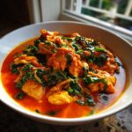 Close-up of a rich, orange-red coconut curry dish featuring chunks of chicken and wilted dark green spinach.