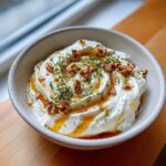 A bowl of creamy whipped ricotta dip swirled and topped with honey, chopped walnuts, and fresh herbs.