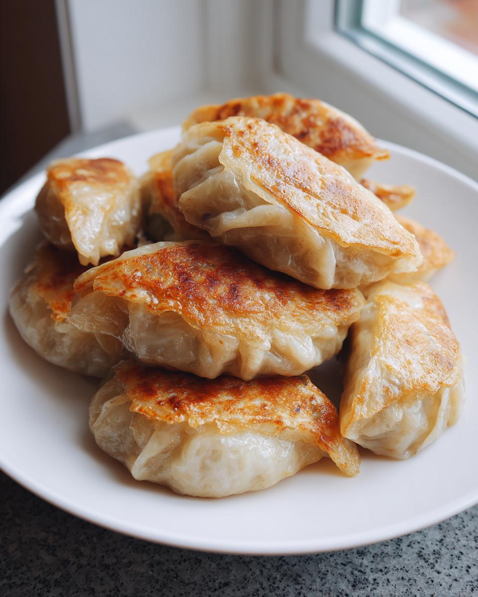 A pile of golden-brown, pan-fried cabbage dumplings stacked on a white plate near a window.