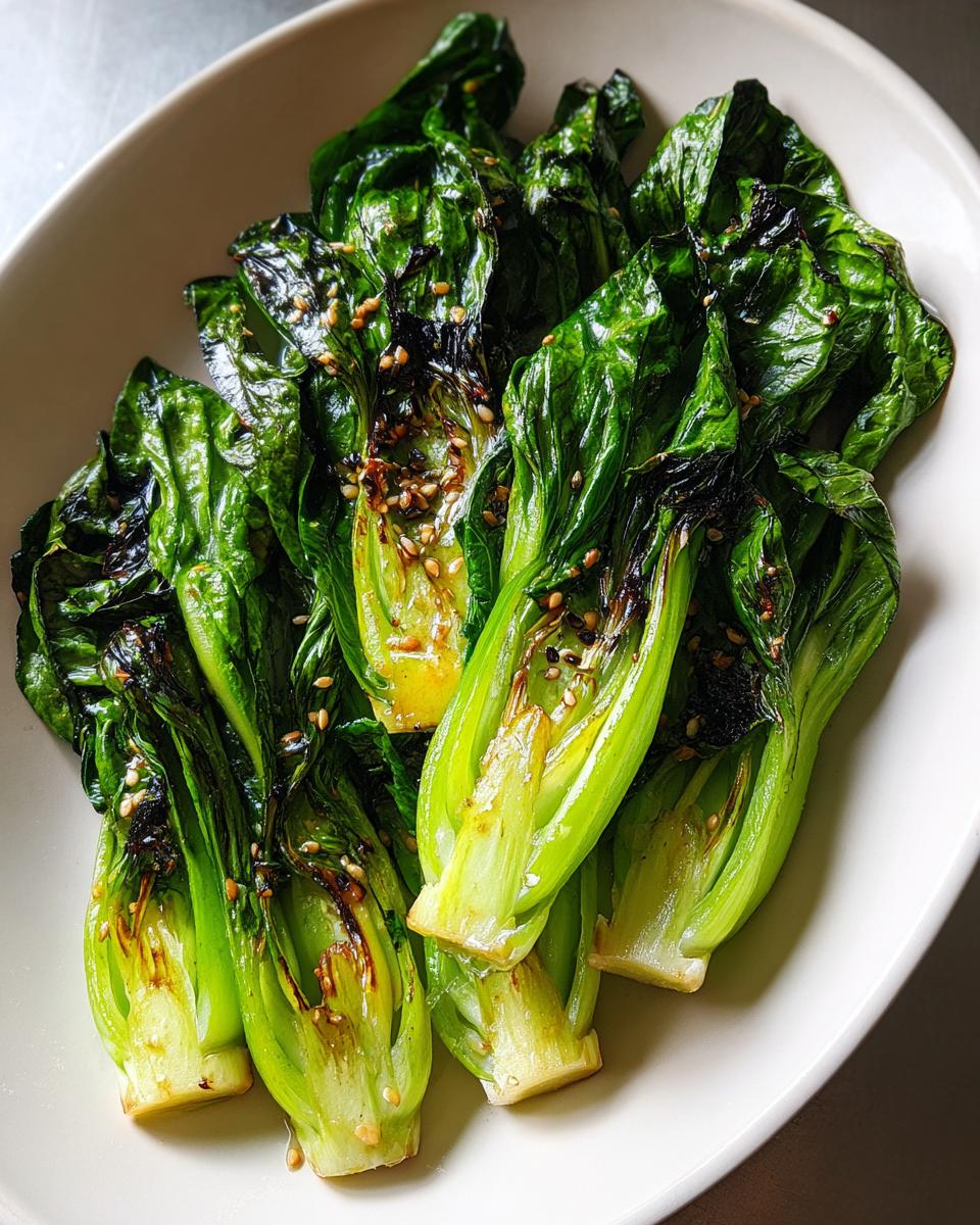 A close-up of vibrant green, glossy sautéed bok choy halves topped with toasted sesame seeds served on a white plate.