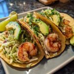 Close-up of three loaded shrimp tacos featuring grilled shrimp, shredded cabbage slaw, avocado slices, and cilantro.