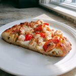 A close-up shot of a single slice of amazing Lobster pizza topped with chunks of lobster meat and cherry tomatoes on a white plate.