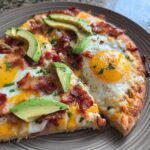 Two slices of a savory breakfast pizza topped with sunny-side-up eggs, crispy bacon, avocado, and cheese.