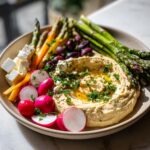 A colorful Mezze platter featuring a large swirl of hummus topped with olive oil and herbs, surrounded by radishes, feta cubes, asparagus, and olives.